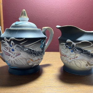 Op de afbeelding: Een zwarte en witte porseleinen suikerpot en roomkan set met een drakenontwerp. De set heeft gouden accenten en blauwe en rode details.