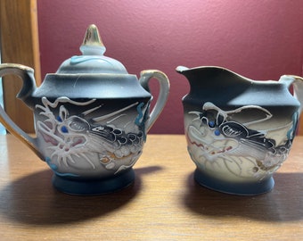 Op de afbeelding: Een zwarte en witte porseleinen suikerpot en roomkan set met een drakenontwerp. De set heeft gouden accenten en blauwe en rode details.