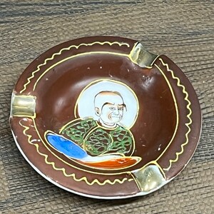 以下が含まれることがあります： 金色の縁取りと緑と青のローブを着た人物の絵が描かれた茶色の陶器製の灰皿。灰皿は波打つ縁取りをしています。