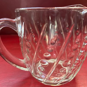 May include: A clear glass jug with a textured swirl design and a handle. The jug has a rounded bottom and a wide mouth.