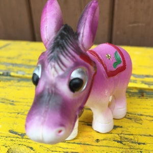 Könnte beinhalten: Eine lilafarbene Plastik-Esel-Figur mit schwarzen Streifen und großen Augen. Der Esel trägt einen rot-grünen Sattel mit einer gelben Blume.
