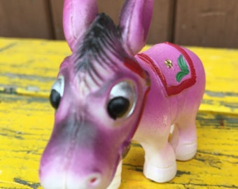Könnte beinhalten: Eine lilafarbene Plastik-Esel-Figur mit schwarzen Streifen und großen Augen. Der Esel trägt einen rot-grünen Sattel mit einer gelben Blume.