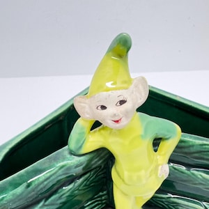 May include: A green and yellow ceramic planter shaped like a fence with a smiling elf leaning on it. The elf is wearing a green and yellow hat and has a green and yellow outfit.