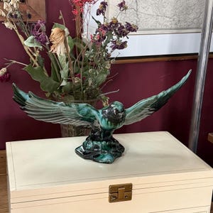 May include: A ceramic eagle sculpture with outstretched wings, in shades of teal, black, and green. The bird is perched on a dark base, with a glossy finish. The sculpture is displayed on a white box with a brass clasp, with dried flowers in the background.