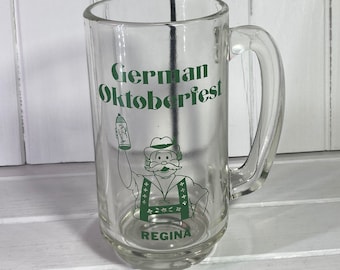 May include: A clear glass beer stein with a green handle. It has a green graphic of a man in traditional German clothing holding a beer stein. The text "German Oktoberfest" and "Regina" are printed on the stein.