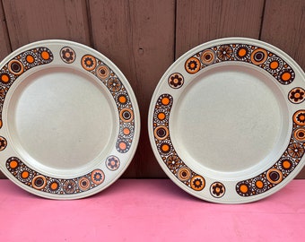 May include: Two white ceramic dinner plates with a brown and orange geometric pattern around the rim.