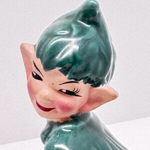 Könnte beinhalten: Eine Keramik-Elfenfigur mit grünen Haaren und einem grünen Outfit. Der Elf zwinkert und hat ein verschmitztes Lächeln.