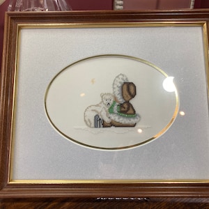 May include: A framed cross-stitch artwork depicting a brown bear wearing a white hooded coat and a white polar bear in a snowy scene.