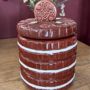 Op de afbeelding: Een keramische koektrommel met een donkerbruine buitenkant en witte accenten. De pot is rond met een deksel en een decoratieve knop. De pot staat op een houten oppervlak.