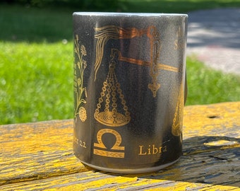 Pode incluir: Caneca de cerâmica preta com um símbolo de Libra do zodíaco dourado e um design de balança. A caneca tem o texto "Sept 22 - Oct 22" e "Libra".