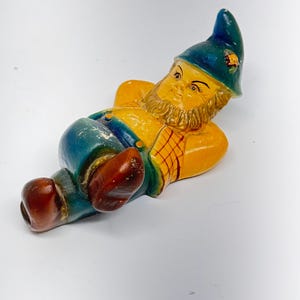 Puede incluir: Una figurita de gnomo de cerámica tumbada de espaldas con los brazos detrás de la cabeza. El gnomo lleva un sombrero azul con una flor, pantalones azules y zapatos marrones. El gnomo tiene un cuerpo amarillo, barba marrón y una cara pintada.