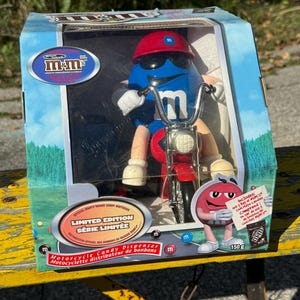 May include: A blue M&M's character wearing sunglasses and a red helmet rides a red motorcycle candy dispenser. The box is labelled "Limited Edition" and "Motorcycle Candy Dispenser".