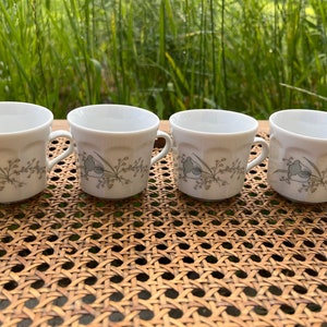 May include: Four white ceramic coffee mugs with a floral design in shades of gray and green. The mugs have a rounded shape and a handle on each side.