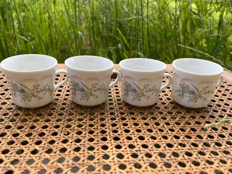 May include: Four white ceramic coffee mugs with a floral design in shades of gray and green. The mugs have a rounded shape and a handle on each side.