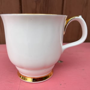 Peut inclure: Une tasse à thé blanche avec un bord et une anse dorés. La tasse a une bande dorée autour du fond.