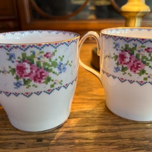 Peut inclure: Deux tasses en céramique blanche avec un motif floral. Les tasses ont un bord doré et un motif floral bleu et rose sur fond blanc.