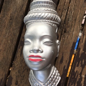 Peut inclure: Sculpture murale en plâtre de couleur argent représentant la tête d'une femme avec une couronne et un collier de perles. Les yeux de la femme sont fermés et elle a des lèvres rouges.