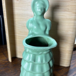 May include: A vintage ceramic planter shaped like a woman in a dress. The planter is a pale green color and has a ruffled skirt. The woman is holding a basket in her arms, which is the planter's opening.