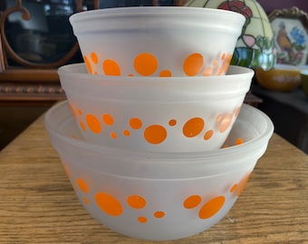 May include: Three white mixing bowls with orange polka dots. The bowls are stacked on top of each other, with the largest bowl on the bottom and the smallest bowl on top.