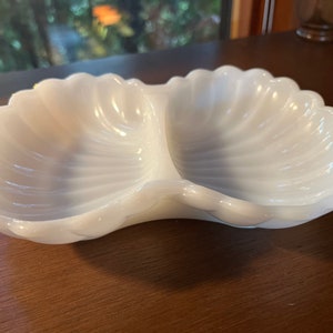 May include: A white ceramic divided dish shaped like two seashells. The dish is made of a smooth, glossy material and has a slightly rippled texture.