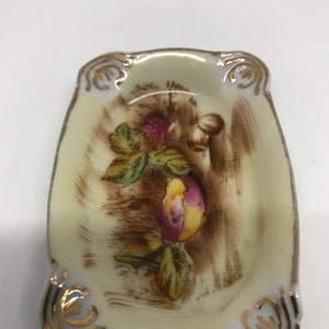 May include: A small, white porcelain dish with a gold rim and a painted design of fruit and leaves. The dish is oval shaped and has a slightly raised edge.