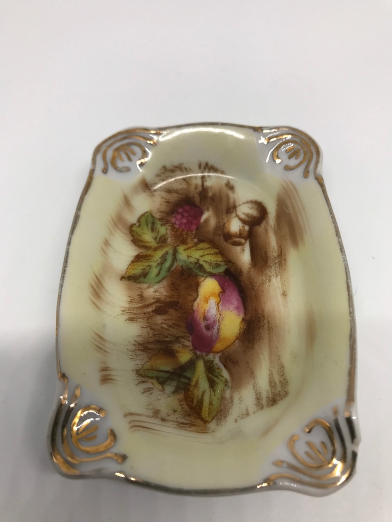 May include: A small, white porcelain dish with a gold rim and a painted design of fruit and leaves. The dish is oval shaped and has a slightly raised edge.