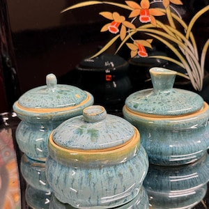 Könnte beinhalten: Drei Keramikgläser mit Deckeln in verschiedenen Größen. Die Gläser haben eine melierte türkisfarbene Farbe mit einem gelb-beigen Rand und Deckelbasis. Die Gläser stehen auf einer reflektierenden Oberfläche.