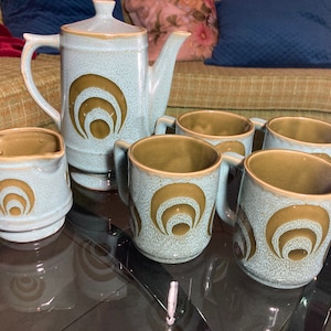 May include: A set of six ceramic mugs and a teapot in a light blue and brown colour scheme. The mugs and teapot feature a concentric circle design in brown on a light blue background.