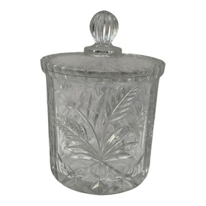 Peut inclure: Un bocal en verre transparent avec un couvercle. Le bocal présente un motif floral et de feuilles gravé dans le verre. Le couvercle a une poignée ronde et nervurée. Le bocal est cylindrique et repose sur un fond blanc.