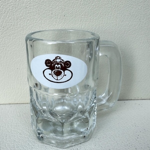 Könnte beinhalten: Klarer Bierkrug aus Glas mit Henkel. Der Becher hat eine rechteckige Form mit strukturiertem Boden. Auf der Vorderseite des Bechers ist ein weißer ovaler Aufkleber mit einem braunen Bärengesicht angebracht.
