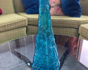 Può includere: Una bottiglia di vetro blu alta con un design testurizzato di uva e viti. La bottiglia ha una base larga e un collo stretto.