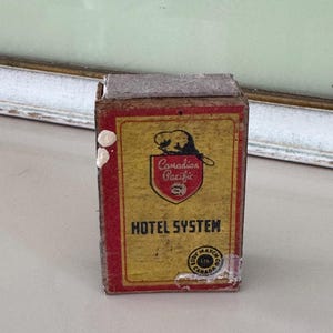 May include: Vintage matchbox with a yellow and red design. The front features the text "Canadian Pacific HOTEL SYSTEM" and a logo with a beaver. The matchbox is slightly worn, showing its age.