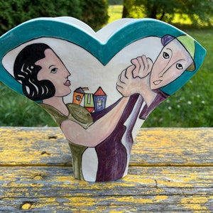Può includere: Un vaso di ceramica bianca a forma di cuore con una scena dipinta di due persone che ballano. La donna indossa un vestito verde e l'uomo indossa una camicia viola e un cappello verde. Lo sfondo è dipinto di colore blu turchese e ci sono piccole case dipinte sullo sfondo.