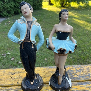 Peut inclure: Une paire de figurines en céramique vintage représentant un danseur et une danseuse. Le danseur est vêtu d'une veste bleue et dorée avec un pantalon noir. La danseuse porte un tutu bleu et noir. Les deux figurines sont debout sur des socles noirs.