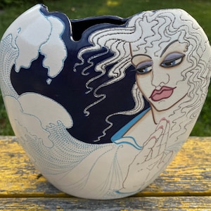 Könnte beinhalten: Eine weiße Keramikvase mit blauem Hintergrund und einem handgemalten Design einer Frau mit wallendem weißem Haar. Das Gesicht der Frau ist mit zarten Zügen bemalt und ihre Hände sind zum Gebet gefaltet. Die Vase ist mit einem blauen und weißen Wellenmuster verziert.