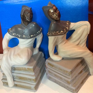 May include: Two ceramic figurines of seated figures atop stacks of books. The figures have bronze-colored faces and wear white robes with gray capes. The books are gray with gold-colored edges. The background is blue.