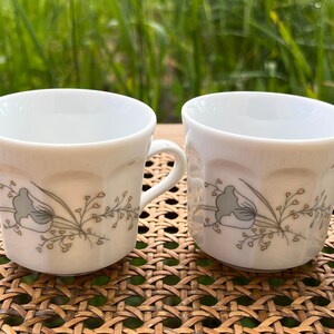 May include: Two white ceramic coffee mugs with a floral design in shades of gray and yellow. The mugs have a fluted design and are sitting on a woven wicker surface.