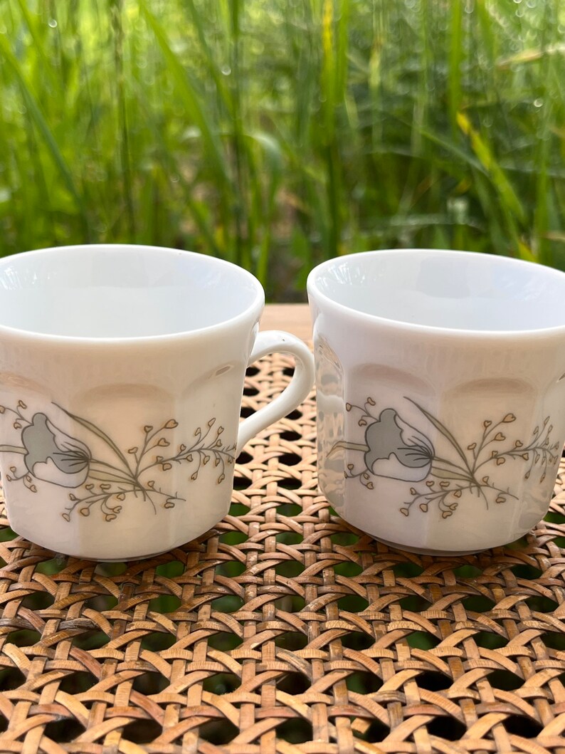 May include: Two white ceramic coffee mugs with a floral design in shades of gray and yellow. The mugs have a fluted design and are sitting on a woven wicker surface.