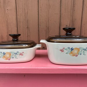 Può includere: Due teglie da forno in ceramica bianca con coperchi in vetro marrone. Le teglie hanno un design floreale con frutta e bacche. Le teglie sono su una superficie rosa.