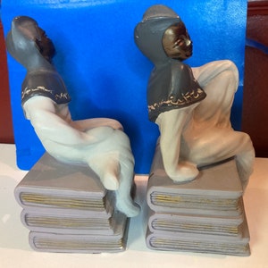 May include: A pair of ceramic bookends featuring figures in a seated pose atop stacks of books. The figures have dark skin, gray hats and capes, and white clothing. The books are gray with gold-colored edges.