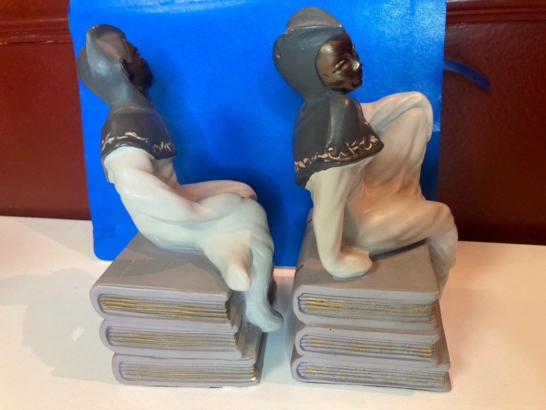 May include: A pair of ceramic bookends featuring figures in a seated pose atop stacks of books. The figures have dark skin, gray hats and capes, and white clothing. The books are gray with gold-colored edges.