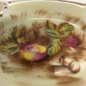 May include: A vintage porcelain ashtray with a hand-painted design of fruit and leaves. The design features a peach, raspberries, and leaves in shades of brown, green, yellow, and pink.