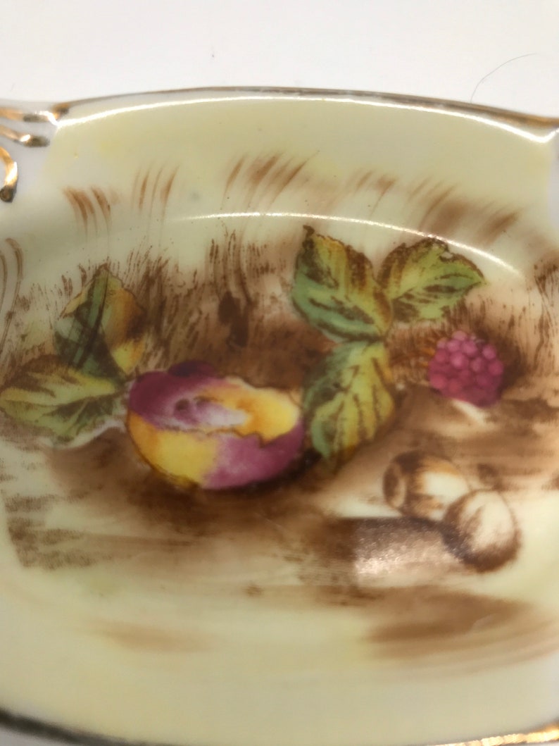 May include: A vintage porcelain ashtray with a hand-painted design of fruit and leaves. The design features a peach, raspberries, and leaves in shades of brown, green, yellow, and pink.