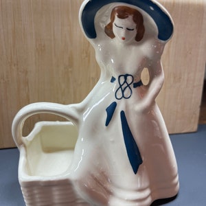 Könnte beinhalten: Ein weißer Keramik-Pflanztopf in Form einer Frau, die einen blau-weißen Hut und ein Kleid trägt. Die Frau hält einen Korb mit einem Griff.