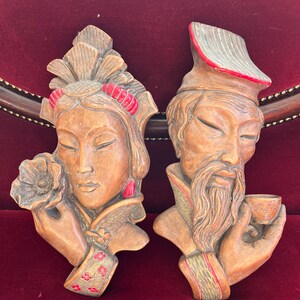 Peut inclure: Une paire de sculptures murales en bois représentant un homme et une femme en vêtements traditionnels asiatiques. La femme tient une fleur et l'homme tient une tasse. Les deux sculptures sont peintes dans des tons de brun, rouge et or.