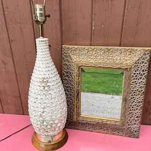 Puede incluir: Una lámpara de mesa de cerámica blanca con un diseño texturizado y puntiagudo y una base dorada. Junto a la lámpara hay un espejo con marco dorado y un diseño ornamentado.
