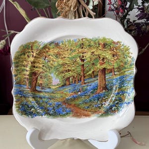 Puede incluir: Plato decorativo con borde festoneado, con una escena de bosque con un sendero, árboles y flores azules. El plato tiene un borde dorado y se exhibe en un soporte blanco. El texto "THIS IS ENGLAND" es visible.
