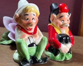 May include: Two ceramic figurines of gnomes sitting with their legs crossed. The gnome on the left is wearing a white hat, red scarf, and green pants. The gnome on the right is wearing a black hat, red and yellow shirt, and black pants. Both gnomes have a white beard and a red nose.