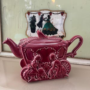 May include: A burgundy-colored ceramic teapot shaped like a carriage, with intricate detailing and a handle. The teapot features a lid with a decorative element and is set against a backdrop of a framed picture with a dancing couple.
