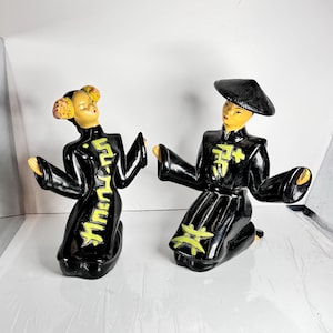 May include: A pair of ceramic figurines depicting a man and woman in traditional Chinese clothing. The man is wearing a black hat and robe with yellow accents, while the woman is wearing a black robe with yellow accents and has her hair styled in two buns. Both figurines are kneeling and have their hands raised in a gesture of greeting.